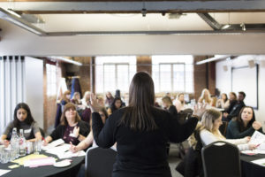 Academy course with facilitator at front with back facing the camera and teachers sitting at tables.