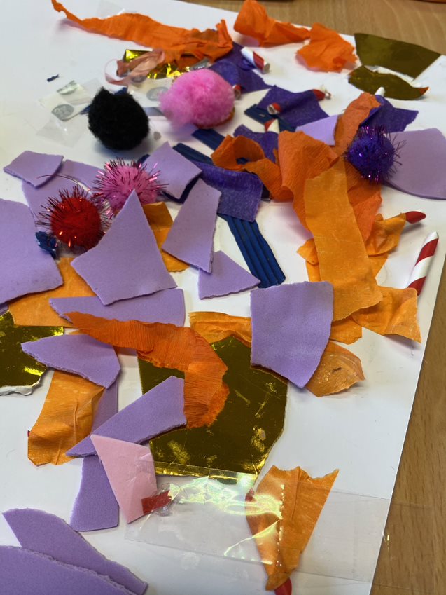 A white board covered with shapes made from purple foam and orange silk, pink and red sparkly pom poms, and gold card. 