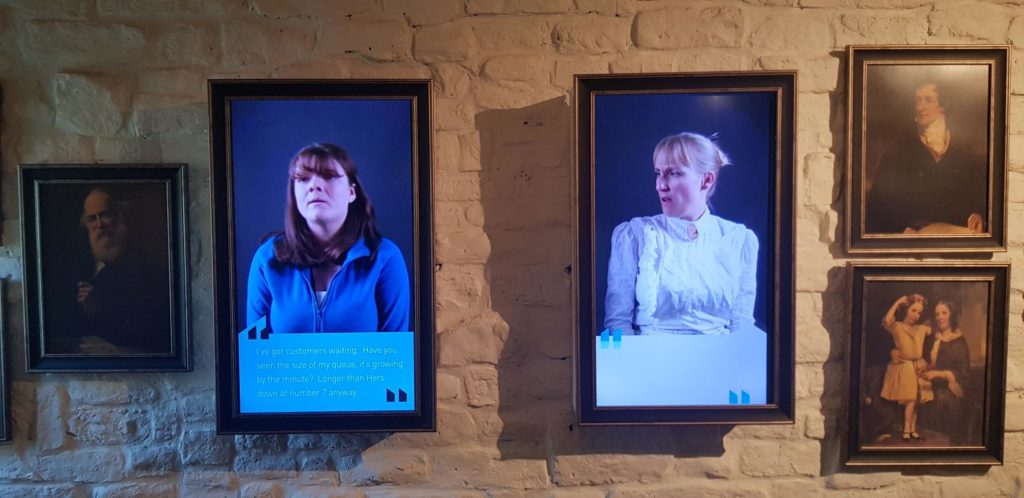 Two screens in wooden picture frames. The frame on the left features the video of a modern shop working wearing blue and the screen on the right features a shop working from the Victorian times wearing a light blue shirt with a white apron and hat