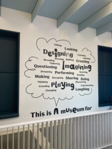 A white wall with black text on. In bold writing reads “This is a museum for”. A giant speech bubble with words such as Designing, Imagining, playing, laughing, thinking inside
