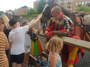 A man in a red outfit with a layer of silver amour on top. He is stood next to a brown horse and children are stroking the horse.