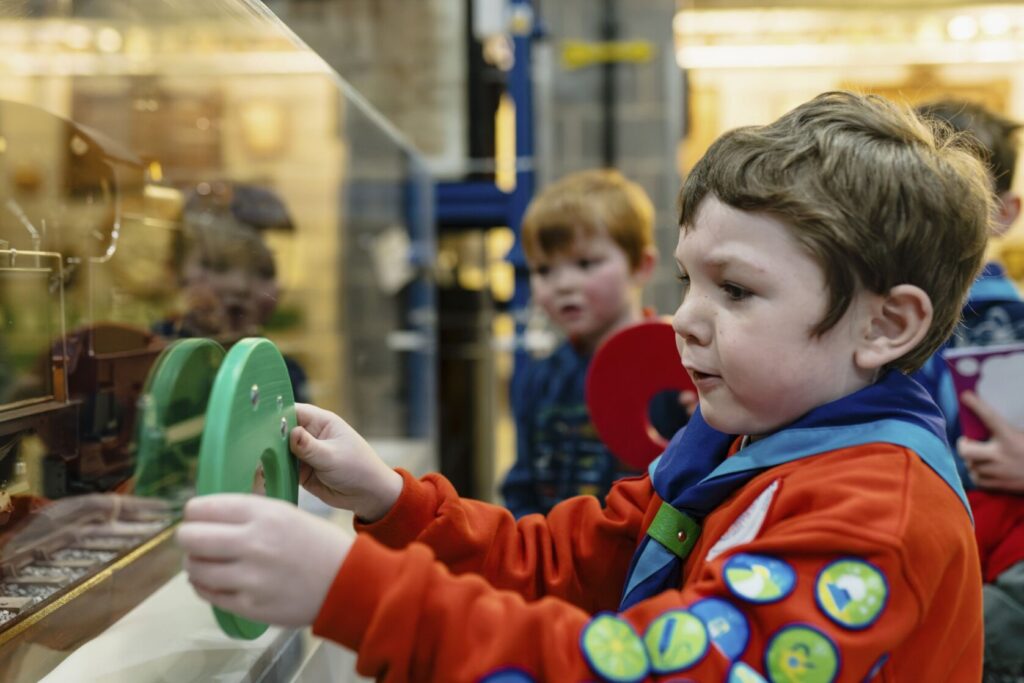 local Squirrel scouts wearing their red jumper and blue neckerchief uniforms hold hungry shaped monsters up against a model train to see if it has any of the same shapes on it. 