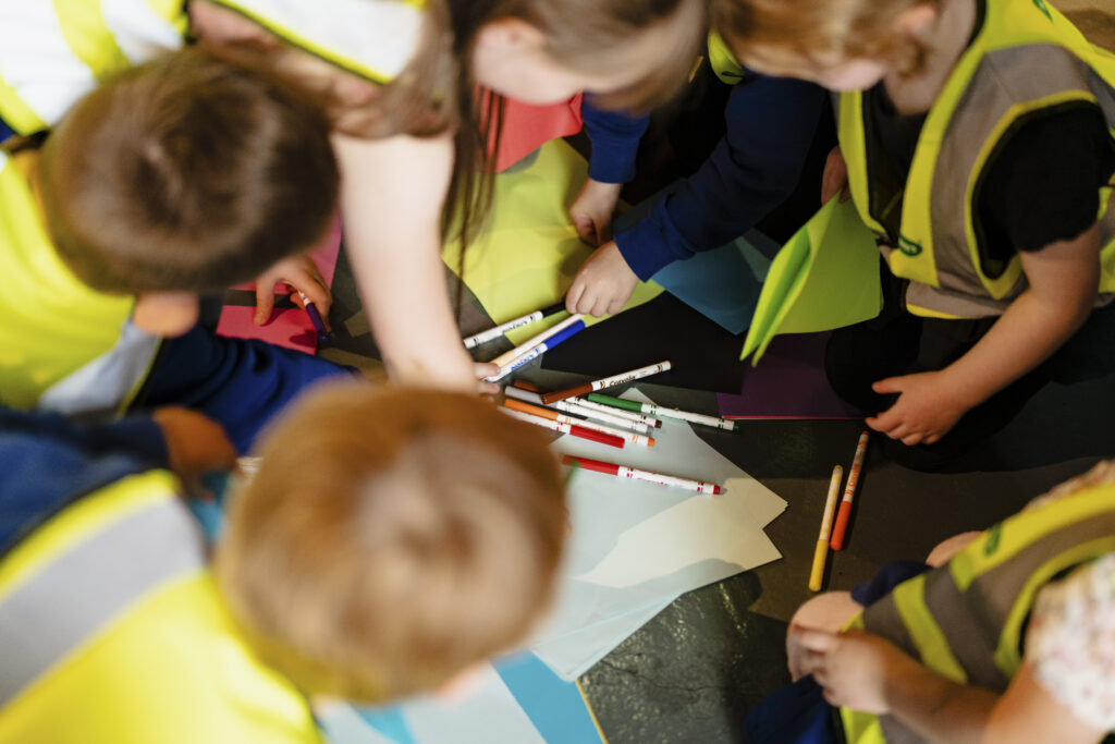 Young children wearing yellow hi vis jackets, are sat on the floor in a circle reaching into the centre to pick up a coloured felt tip pen. 