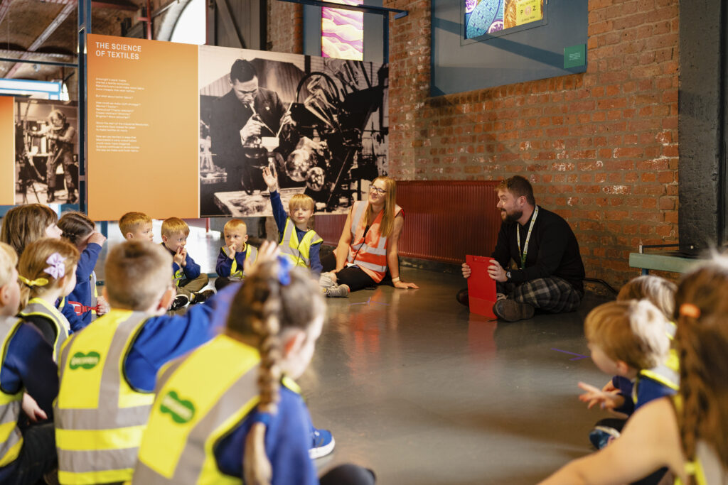 A class of young children with their teacher sit in a semi circle all wearing hi vis jackets, some of the children have their hands up to answer a question asked by a museum employee sat in the middle. 